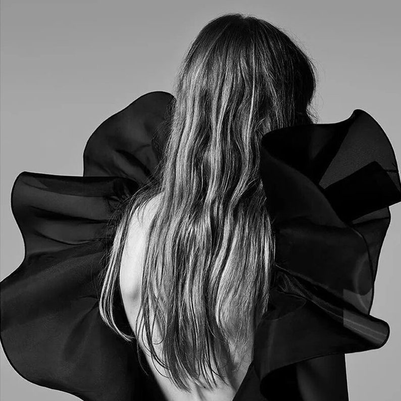 May include: A black and white photo of a person with long hair wearing a black ruffled garment. The person is facing away from the camera.