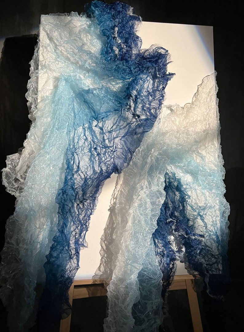 Puede incluir: Una pieza de arte abstracto azul y blanco hecha de tela. La tela est&aacute; drapeada sobre un lienzo y parece estar flotando en el viento.