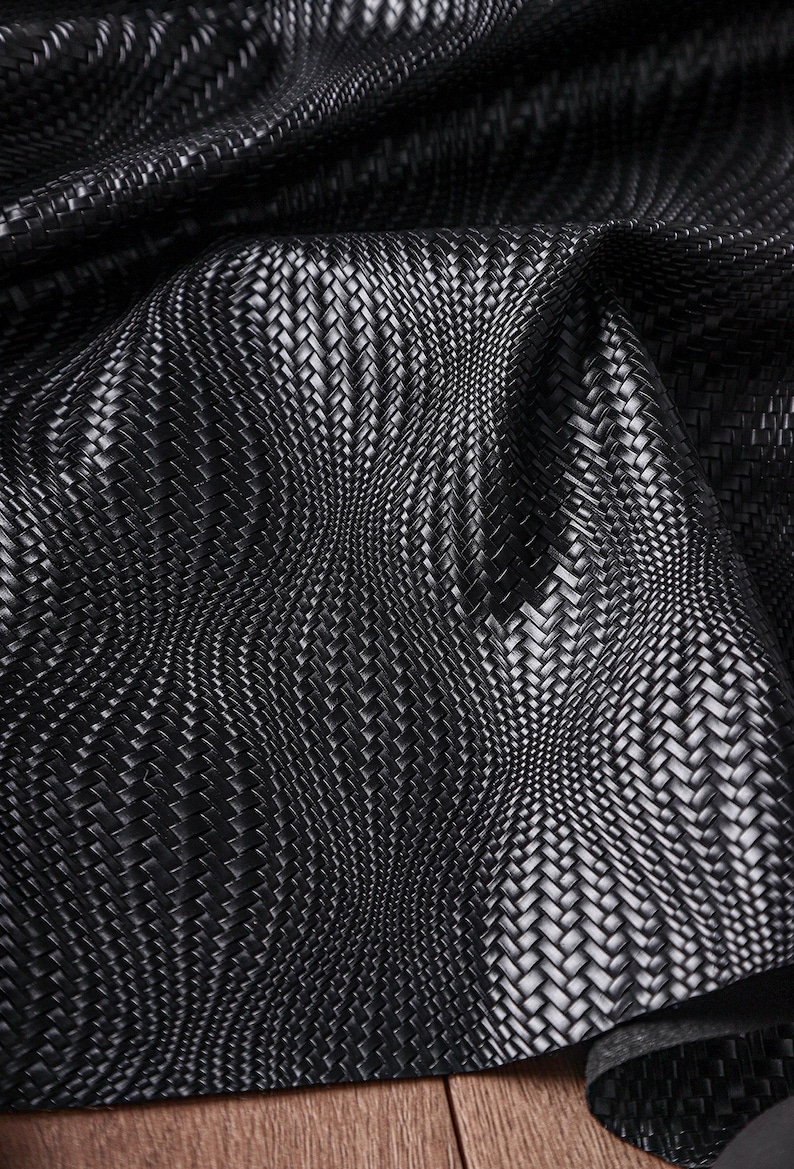 May include: Close-up of a black woven fabric with a textured basketweave pattern. The material has a subtle sheen, with light reflecting off the raised areas. The fabric is draped, creating soft curves and shadows.