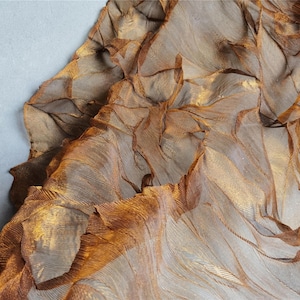 May include: Close-up of a textured, layered fabric in shades of brown and gold. The material appears to be sheer, with a rippled, organic design. The fabric is draped over a light gray surface.