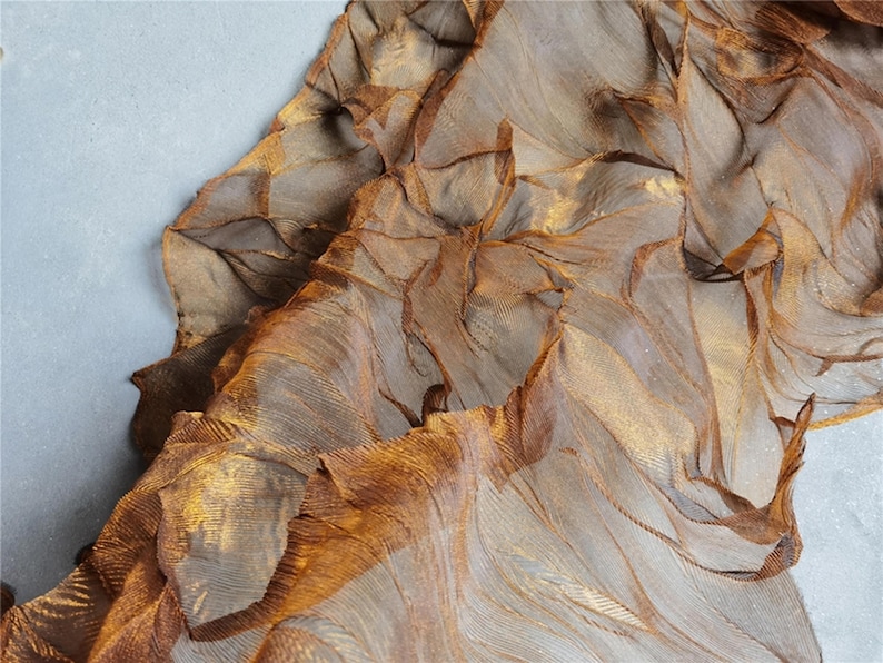 May include: Close-up of a textured, layered fabric in shades of brown and gold. The material appears to be sheer, with a rippled, organic design. The fabric is draped over a light gray surface.