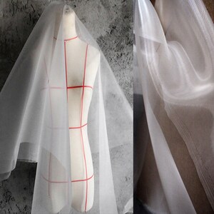 May include: White sheer tulle fabric with a fine mesh texture. The fabric is draped over a dress form and shown in a close-up.