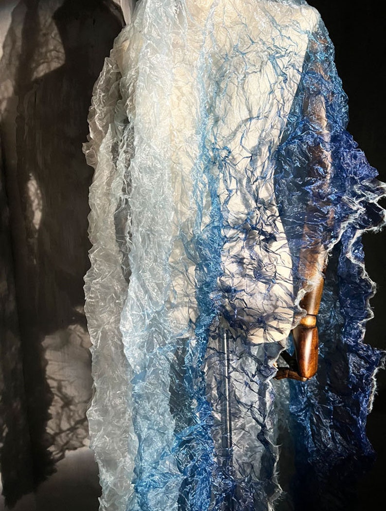 Puede incluir: Una bufanda ligera y transparente con un degradado de colores azul y blanco. La bufanda est&aacute; hecha de una tela delicada y arrugada y tiene una apariencia fluida y et&eacute;rea.