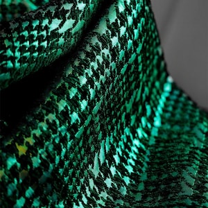 May include: Close-up of a folded fabric with a houndstooth pattern. The fabric is a deep teal green with black accents, creating a textured appearance. The folds create shadows, highlighting the pattern and color variations.