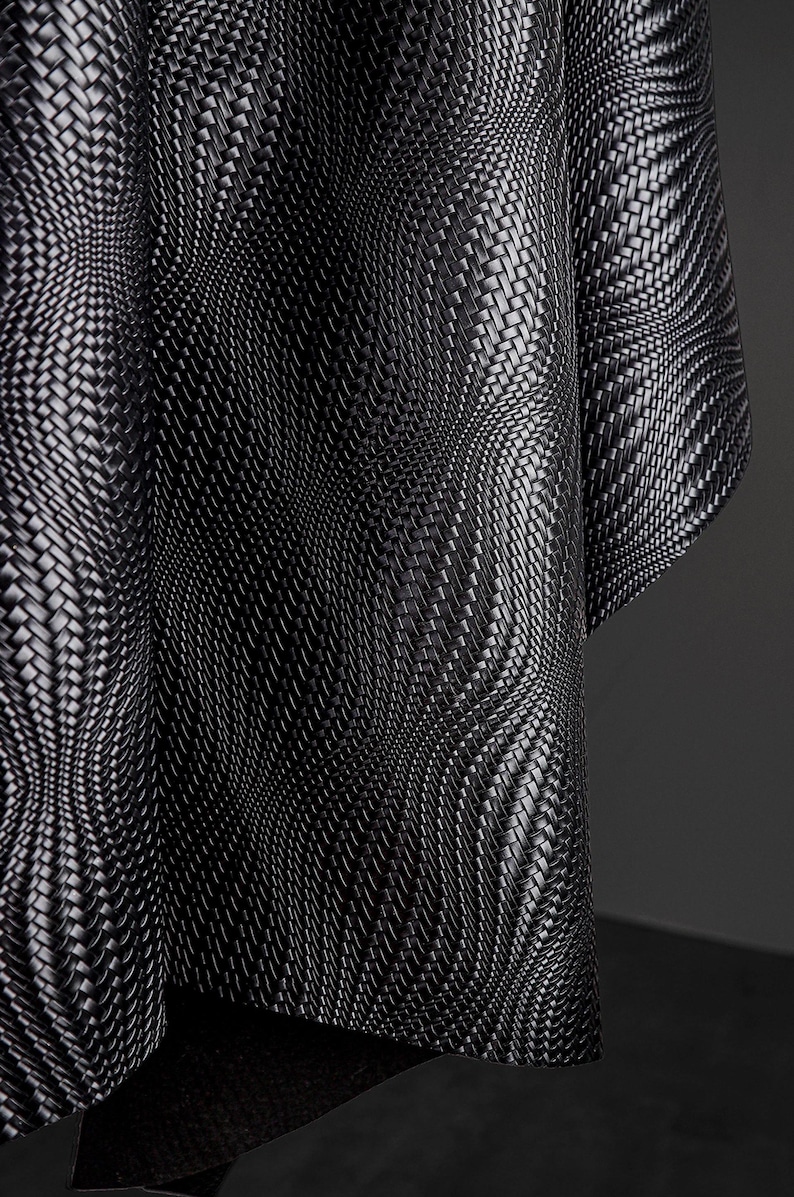 May include: Close-up of a black woven fabric with a textured surface. The fabric is draped, creating curved lines and shadows. The weave pattern is intricate, with a repeating design. The overall effect is a modern, abstract textile.