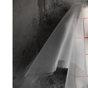 May include: A sheer white fabric draped over a textured gray surface. The fabric is translucent and has a slight sheen.
