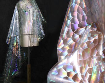 May include: Iridescent, clear fabric with a honeycomb pattern draped over a dress form. The fabric reflects a rainbow of colors, creating a shimmering effect. The dress form is white, and the background is black.