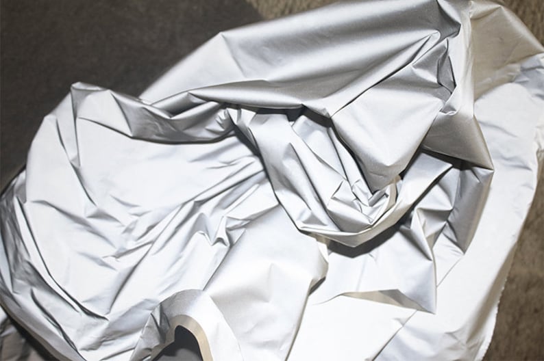 May include: A silver reflective fabric with a smooth, shiny surface. The fabric is crumpled and folded, showing its texture.