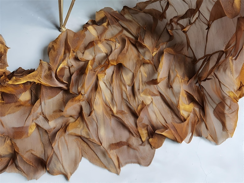 May include: A flowing, decorative fabric in shades of brown and gold, with a layered, textured appearance. The fabric appears to be draped, with a light, airy quality. The material is likely silk or a similar delicate textile.