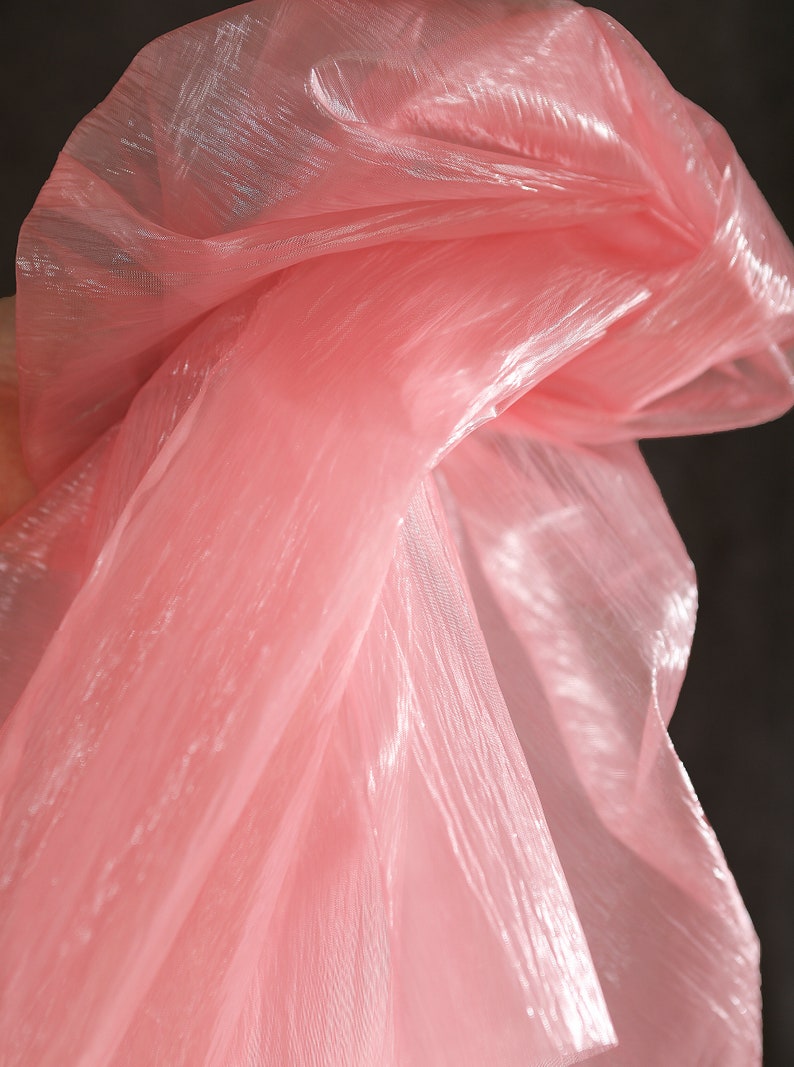 May include: Pink sheer fabric with a shimmery finish. The fabric is draped and folded, creating soft folds and wrinkles.