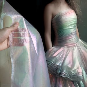 May include: Iridescent fabric held up next to a strapless gown. The fabric and gown have a shimmering, multi-colored effect with hues of pink, green, and blue. The gown features a layered skirt.
