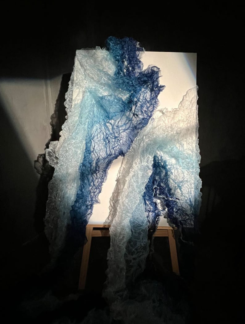 Puede incluir: Una escultura de tela azul y blanca sobre un caballete de madera. La tela est&aacute; drapeada y fluye, creando una sensaci&oacute;n de movimiento y fluidez.