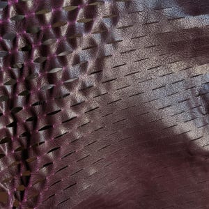Puede incluir: Un material de color morado oscuro con una superficie texturizada y granulada que presenta un patr&oacute;n repetitivo de ranuras horizontales cortadas con l&aacute;ser. Los bordes de los cortes revelan una capa interior de color magenta brillante.
