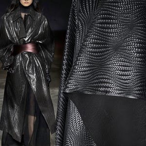 May include: A black, textured coat with a wrap design and a wide burgundy belt. The coat has a shiny, woven appearance. The image also shows a close-up of the fabric's intricate pattern, which is a repeating weave in shades of black and grey.