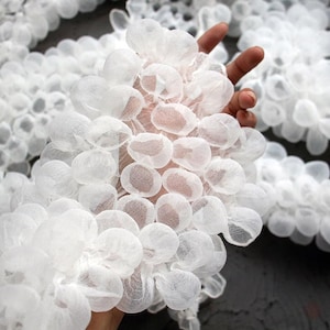 Op de afbeelding: Een close-up van een witte, organisch ogende textiel. Het textiel bestaat uit talrijke kleine, afgeronde, bloembladachtige vormen, waardoor een delicate, wolkenachtige uitstraling ontstaat. De textuur is zacht en luchtig, met een licht doorschijnende kwaliteit.