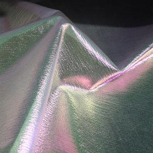 May include: Iridescent fabric with a textured surface. The fabric has a color-shifting effect, displaying hues of green, pink, and silver. The material is folded, creating sharp angles and highlighting the play of light across its surface.