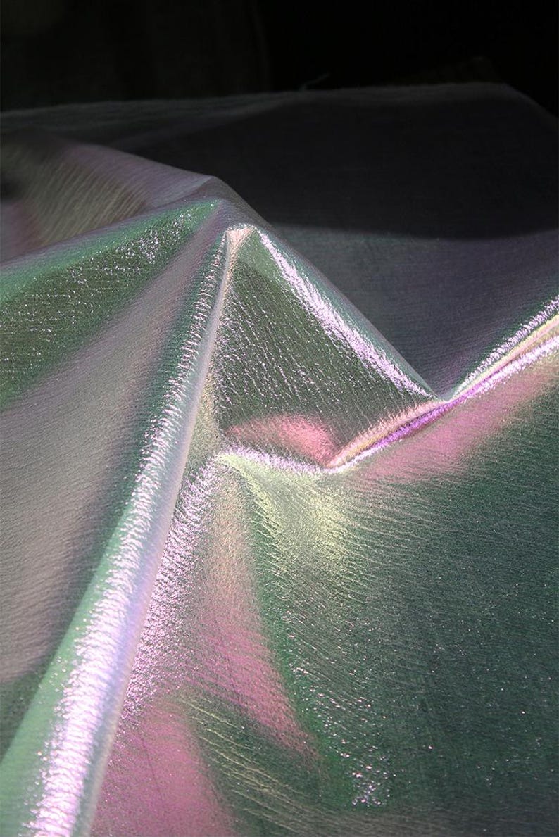 May include: Iridescent fabric with a textured surface. The fabric has a color-shifting effect, displaying hues of green, pink, and silver. The material is folded, creating sharp angles and highlighting the play of light across its surface.
