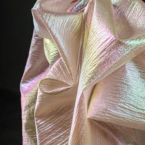 May include: Close-up of iridescent fabric with a textured surface. The fabric is folded and draped, showcasing a play of colors including pink, green, and yellow. The material has a shimmering, pearlescent quality.
