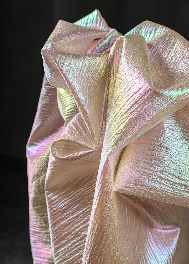 May include: Close-up of iridescent fabric with a textured surface. The fabric is folded and draped, showcasing a play of colors including pink, green, and yellow. The material has a shimmering, pearlescent quality.