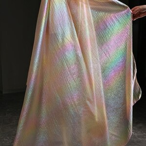 May include: Iridescent fabric with a textured surface. The fabric displays a spectrum of colors, including pink, yellow, green, and blue, creating a shimmering effect. The material is draped, showcasing its flowing quality.