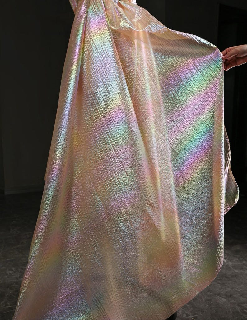 May include: Iridescent fabric with a textured surface. The fabric displays a spectrum of colors, including pink, yellow, green, and blue, creating a shimmering effect. The material is draped, showcasing its flowing quality.
