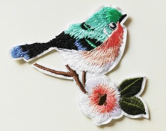 Puede incluir: Parche bordado para planchar con un pájaro colorido posado en una rama con una flor rosa. El pájaro tiene un cuerpo verde, azul y rosa con alas negras. La flor tiene pétalos blancos y un centro rosa. El parche está etiquetado como "TYPE A".