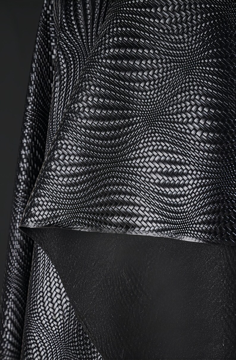 May include: Close-up of a black and white woven fabric with a textured pattern. The fabric appears to be layered, with a contrasting black material visible underneath. The weave creates a dynamic, three-dimensional effect.