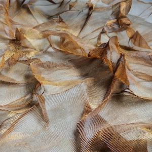 May include: Close-up of a textured fabric with a layered, rippled effect. The fabric has a metallic sheen with shades of gold and bronze. The material appears to be lightweight and flowing, with visible lines and folds creating depth and dimension.