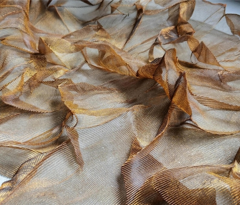 May include: Close-up of a textured fabric with a layered, rippled effect. The fabric has a metallic sheen with shades of gold and bronze. The material appears to be lightweight and flowing, with visible lines and folds creating depth and dimension.