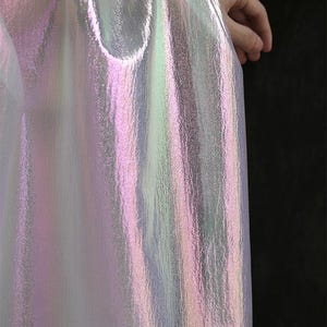 May include: Iridescent fabric with a pearlescent sheen, displaying hues of pink, green, and silver. The material has a textured surface and is draped, creating soft folds. The fabric is held by a hand, with a dark background.