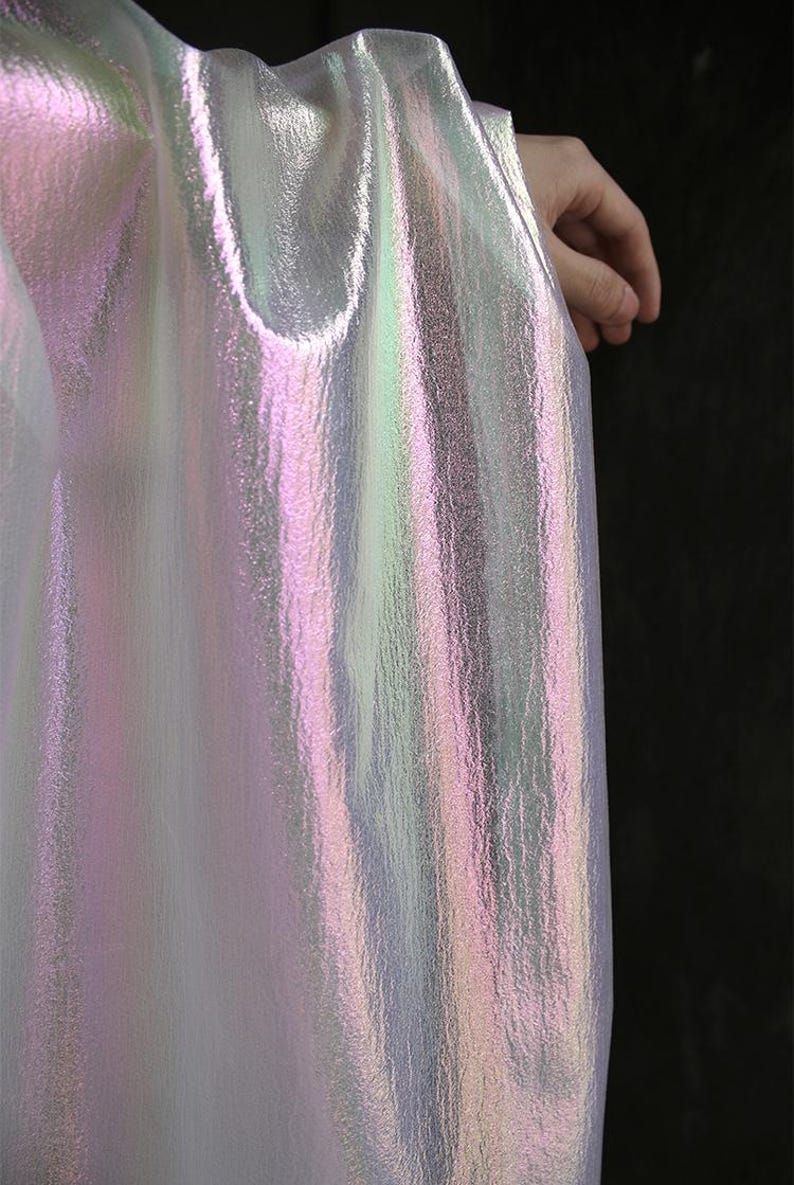 May include: Iridescent fabric with a pearlescent sheen, displaying hues of pink, green, and silver. The material has a textured surface and is draped, creating soft folds. The fabric is held by a hand, with a dark background.