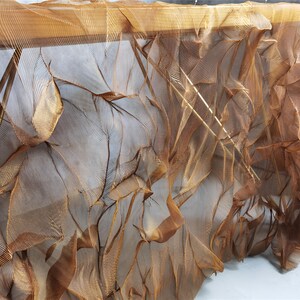 May include: A close-up of a textured, draped fabric in shades of brown and gold. The fabric appears to be layered, with a delicate, net-like structure. The folds and shadows create a sense of depth and movement.