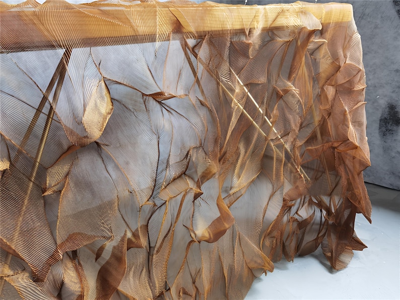 May include: A close-up of a textured, draped fabric in shades of brown and gold. The fabric appears to be layered, with a delicate, net-like structure. The folds and shadows create a sense of depth and movement.