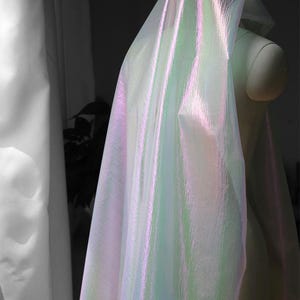May include: Iridescent fabric draped over a mannequin, showcasing a shimmering effect. The fabric transitions between shades of pink, green, and white, creating a luminous appearance. The material appears lightweight and flowing, suggesting its use in fashion or decorative applications.