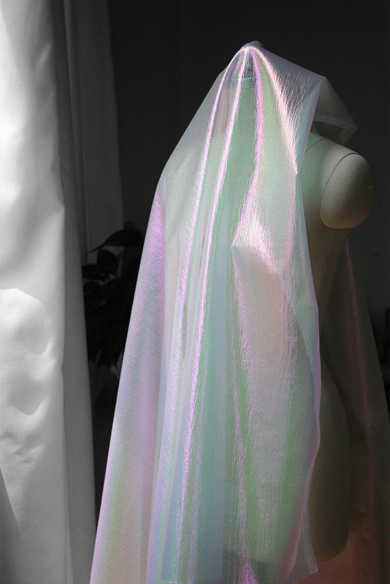 May include: Iridescent fabric draped over a mannequin, showcasing a shimmering effect. The fabric transitions between shades of pink, green, and white, creating a luminous appearance. The material appears lightweight and flowing, suggesting its use in fashion or decorative applications.