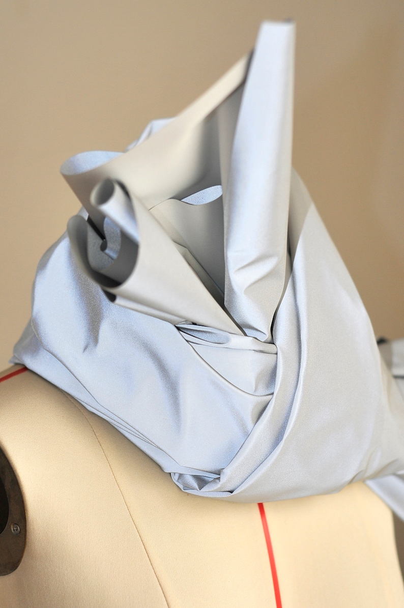 May include: A silver reflective fabric draped over a dress form. The fabric is folded and bunched up, creating a soft, flowing look.