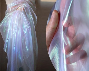 May include: Iridescent fabric draped over a mannequin, showcasing a shimmering, translucent material. The fabric has a soft, flowing texture with a subtle color shift, reflecting hues of pink, blue, and green. A hand gently holds a section of the fabric, highlighting its delicate nature.