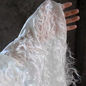 Könnte beinhalten: Ein durchscheinender, weißer Stoff mit einem wellenförmigen, strukturierten Muster. Der Stoff wird hochgehalten, wodurch seine transparente Qualität sichtbar wird. Die Kanten haben einen Fransenbesatz, der zu seinem ätherischen Aussehen beiträgt.