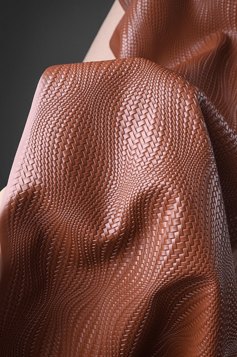 May include: A close-up shot of brown leather fabric with a basketweave design. The material is folded, revealing its texture and depth. The colour is a warm, reddish-brown, similar to tan.