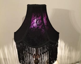 May include: A bell-shaped lampshade featuring a black lace overlay with a floral pattern, revealing a vibrant purple interior light. The bottom edge is finished with long, black hanging fringe trim that extends downward from the scalloped base.