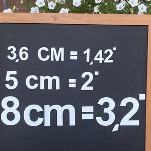 Może przedstawiać: Czarna tablica w drewnianej ramie z białym napisem przedstawiającym przeliczniki miar: 3,6 cm = 1,42 cala, 5 cm = 2 cale i 8 cm = 3,2 cala. Tablica znajduje się przed białymi kwiatami.