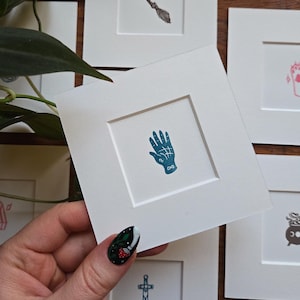 May include: A selection of small, framed art prints with mystical and witchy symbols. The central print shows a blue palmistry hand. Other prints depict a broom, a cauldron, a dagger, and a paw print. The prints are on white paper.