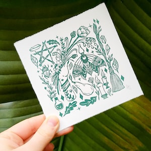 Può includere: Una stampa quadrata bianca con una dettagliata illustrazione verde di una falena, elementi botanici e simboli mistici. L'opera d'arte include un pentagramma, bottiglie di pozioni, candele e una scopa, con una mano che tiene un mazzo di fiori.