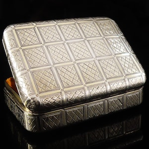 May include: A silver rectangular box with a hinged lid, partially open. The box has a repeating geometric pattern of squares and diagonal lines. The interior of the box is a warm gold colour. The box is on a black surface.