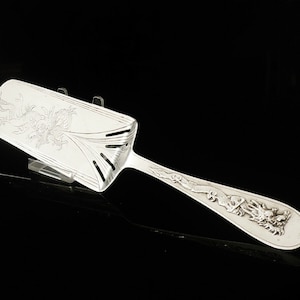 May include: A silver serving spoon with an ornate design featuring a dragon and floral motifs. The spoon has a long handle and a wide, flat bowl.