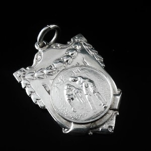 May include: Silver pendant with a shield-shaped design. The front features a circular image of two figures in a wrestling match. The pendant has a loop at the top for hanging.