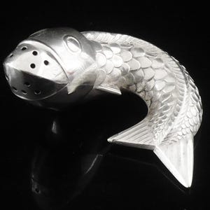 May include: A silver fish-shaped salt or pepper shaker. The shaker has a detailed scale pattern on its body and a tail fin. The head features holes for dispensing contents. The object is set against a black background.