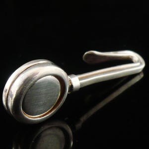 May include: A silver-toned metal hook with an oval-shaped head and a curved handle. The hook is set against a black background, with a reflection visible below.
