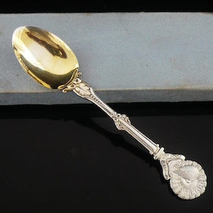 May include: A silver spoon with a gold-coloured bowl, resting on a light blue box. The spoon has ornate detailing on the handle and a decorative end. The spoon is likely a vintage or antique item.