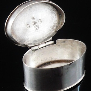 May include: A silver oval-shaped pill box with a hinged lid. The inside of the lid is marked with the letters "SP" and a crown.